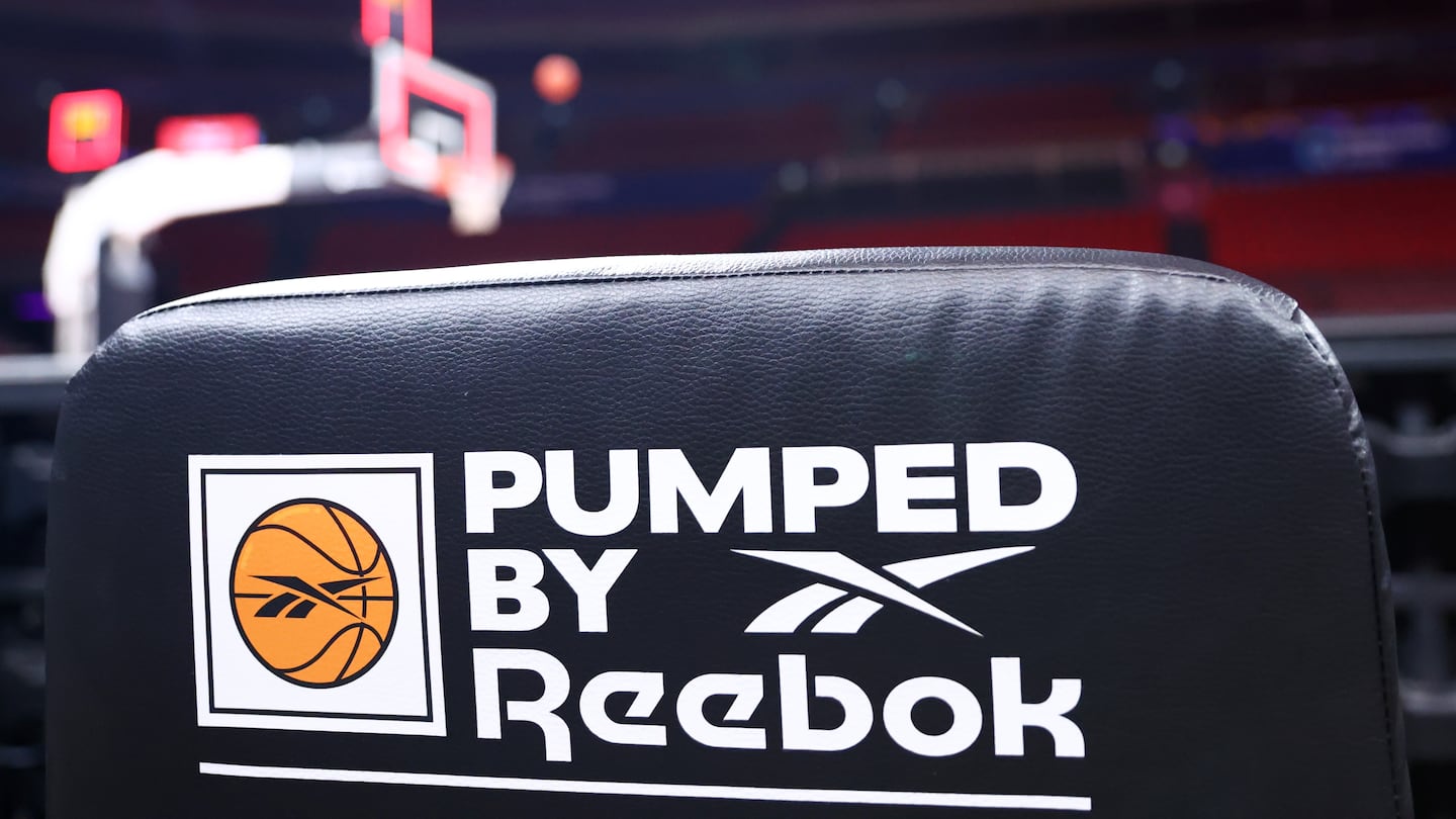 The back of a chair on a basketball court that reads "Pumped by Reebok" with Reebok's logo inside of a basketball inside of a box on the left side.